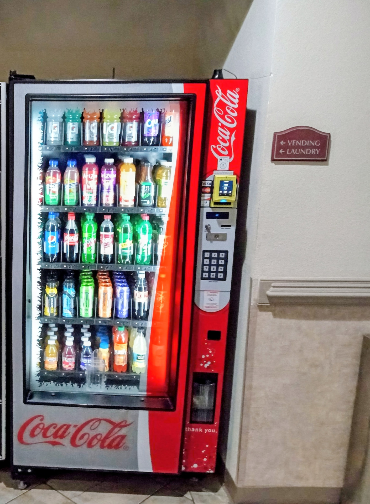 Glass front Coca-Cola soda vending machine stocked with sodas, sports drinks, energy drinks, and bottled water, with a card reader for tap-to-pay
